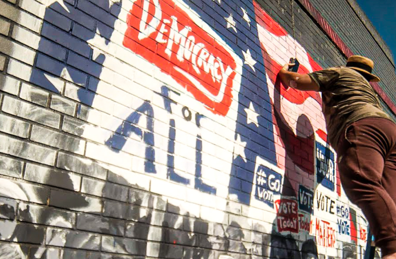 Painter on a ladder works on a mural that depicts protestors with signs reading "Democracy for All"