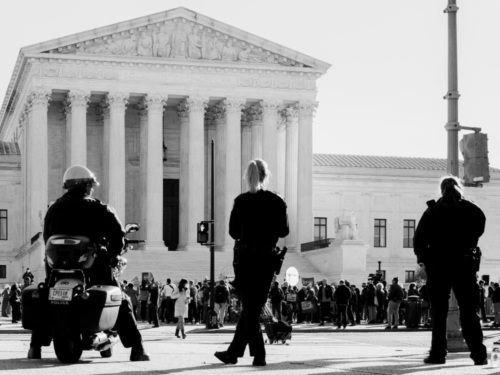 Three police officers keep watch on a crowd of protestors in front of the Supreme Court.