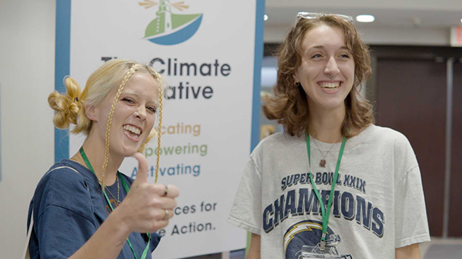 Jules and Maggie smile in front of a sign at a climate conference in 2023. Jules gives the camera a thumbs up.