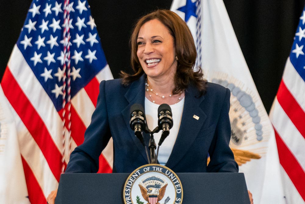 Vice President Kamala Harris smiles at a podium with American flags and vice presidential seal flag in background.