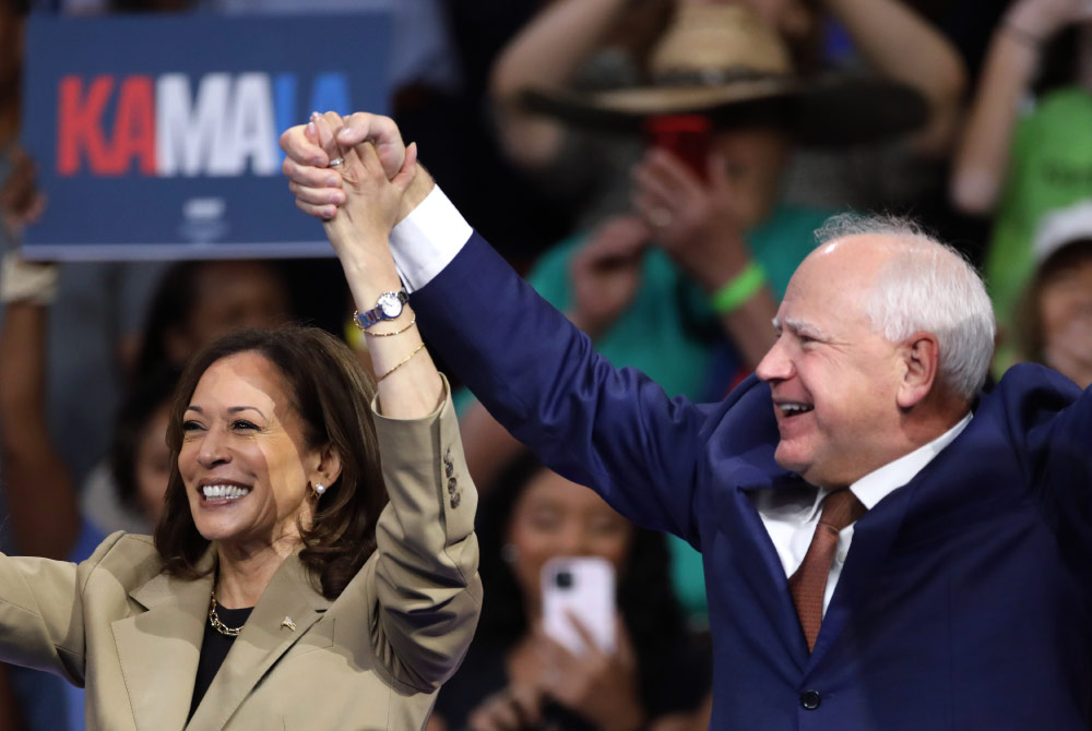 Vice President Kamala Harris and Governor Tim Walz smile and raise hands together on stage at a campaign rally