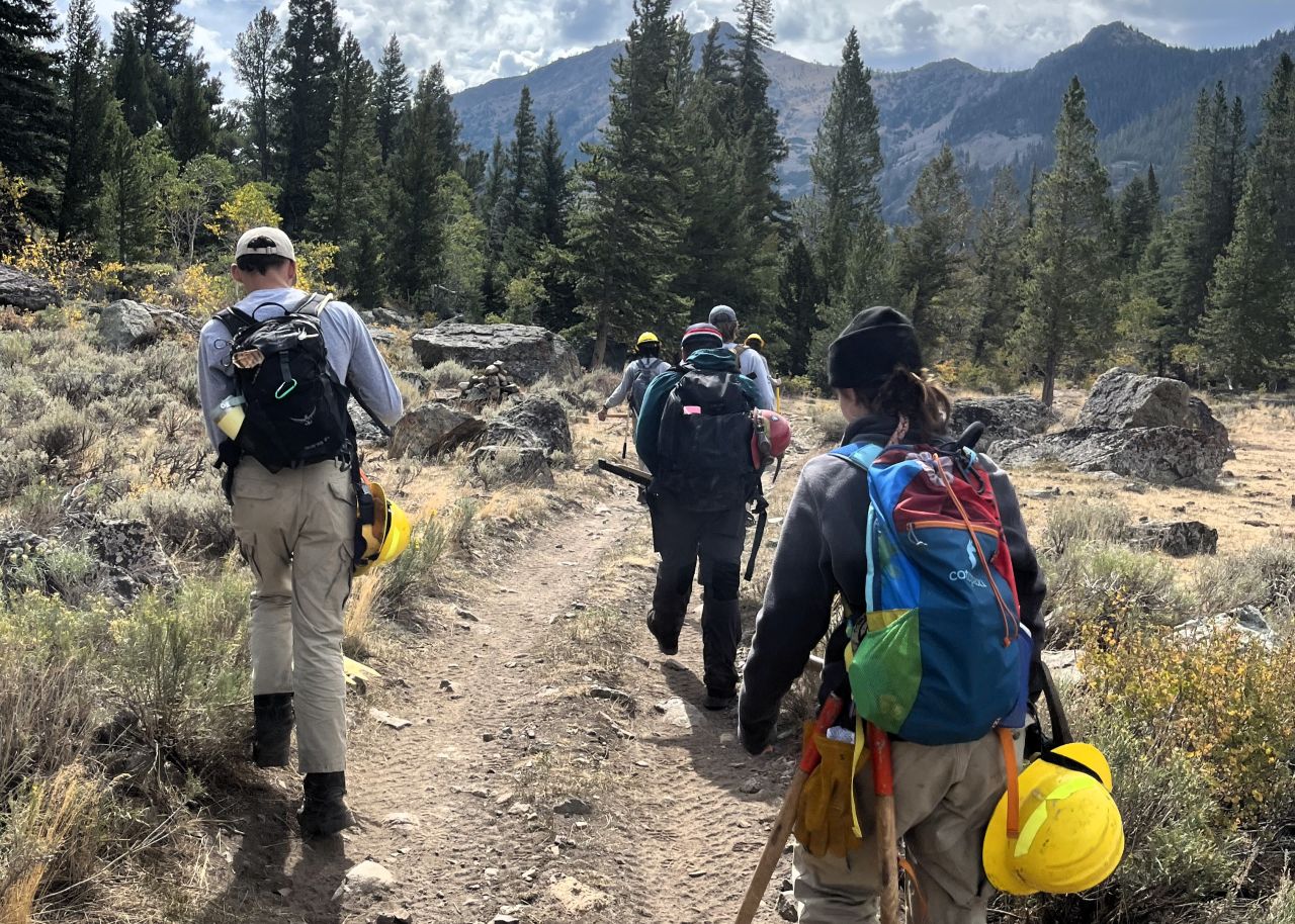 A group of AmeriCorps volunteers carrying backpacks and equipment walk on a dirt path against a backdrop of mountains and trees.