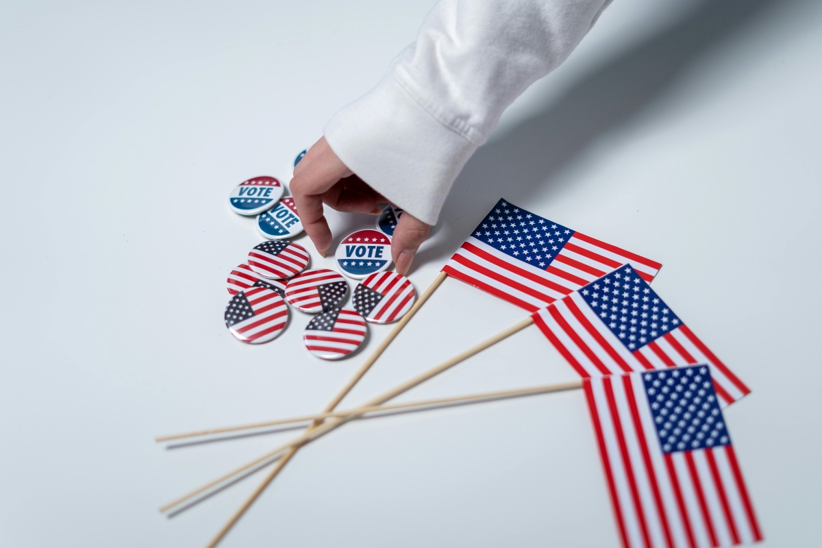 A hand reaching for a pile of pins with the American flag or "Vote" on them, next to three small American flags