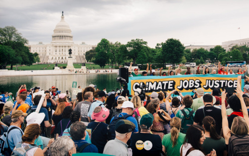 A crowd of Earth Day marchers with a sign reading "Climate, Jobs, Justice" and the Capitol building visible in the background.