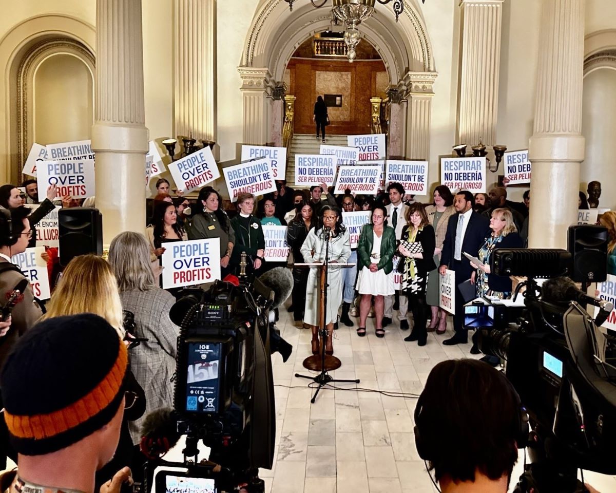 A crowd holding signs that read "People over Profits" and "Breathing shouldn't be dangerous" gathers around a speaker at a podium inside the Colorado State Capitol.